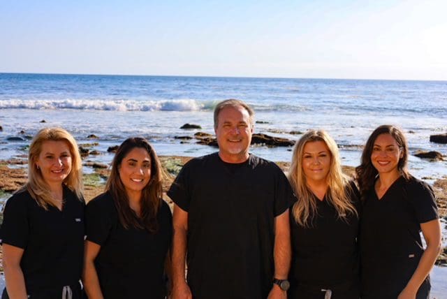 Group of five people smiling at the beach with waves in the background.