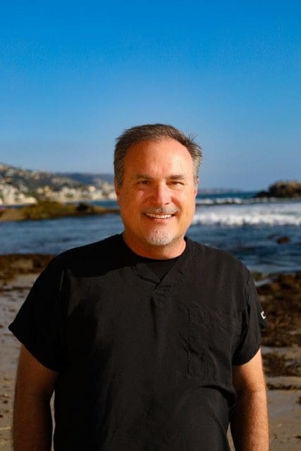 Smiling man in a black shirt by the ocean on a sunny day.