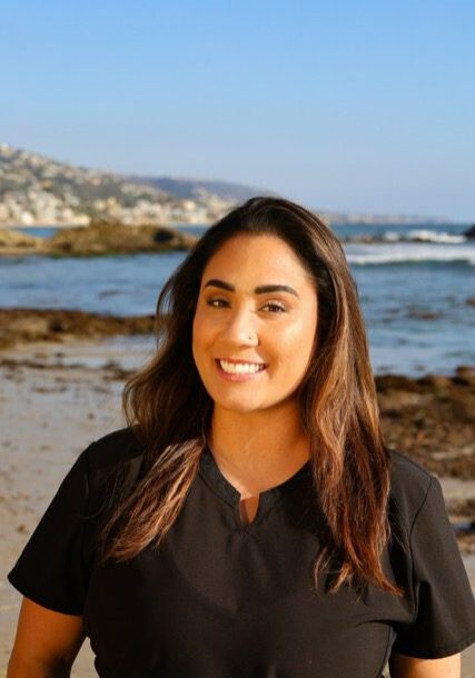 Smiling woman stands by the ocean with coastal hills in the background.